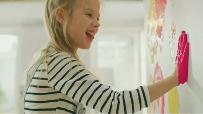 双手蘸黄色和粉红色油漆的快乐小女孩在墙上贴了五颜六色的手印。她玩得很开心，笑得很开心。家正在装修。
