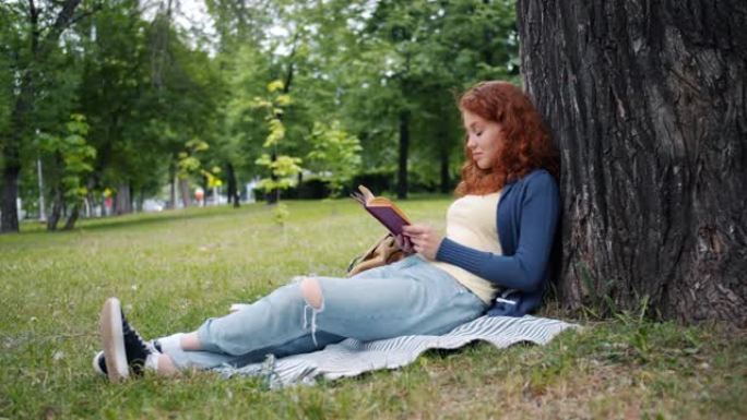 美丽的年轻女子看书坐在公园树下的毯子上微笑