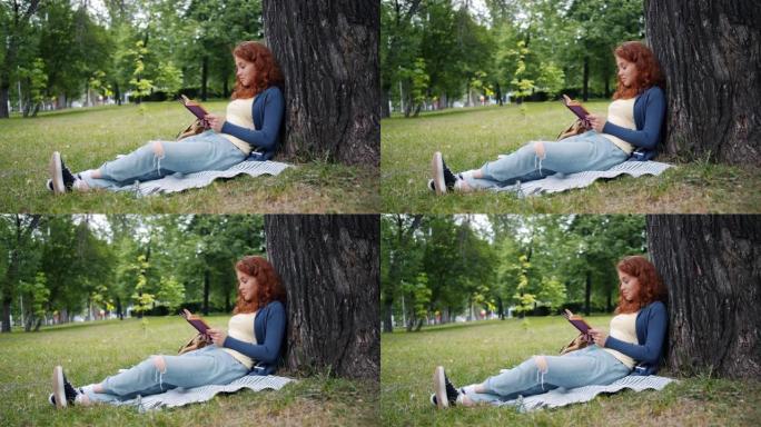 美丽的年轻女子看书坐在公园树下的毯子上微笑