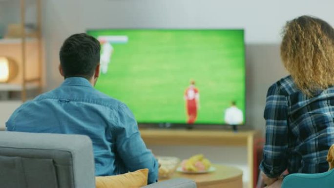 在家里，年轻夫妇在电视上观看足球比赛，体育迷在情感上为他的球队打手势，忧虑和欢呼。倒车摄像头拍摄。