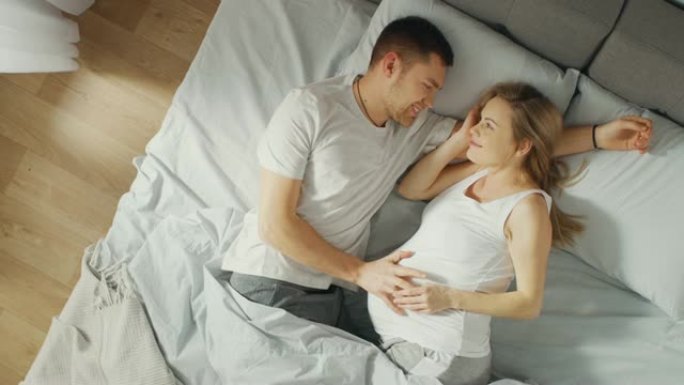 幸福的年轻夫妇在床上拥抱在一起，年轻的女人怀孕了，充满爱心的未来父亲温柔地抚摸着她的肚子。自上而下的