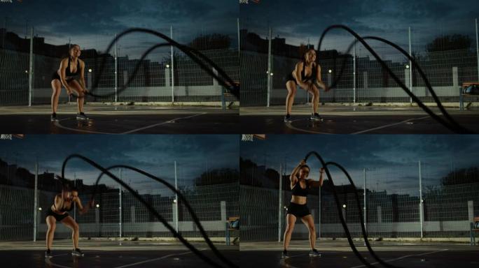 美丽精力充沛的健身女孩用战斗绳锻炼。她正在一个有围栏的室外篮球场里锻炼身体。居民区下雨后的晚间录像。