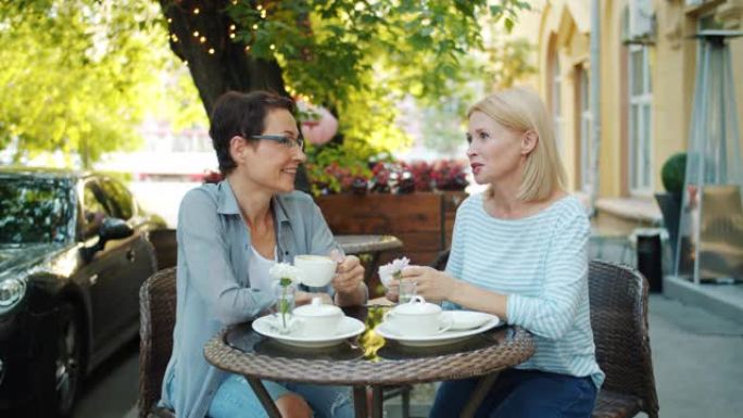 迷人的女士朋友在户外咖啡馆聊天，手里拿着咖啡杯微笑