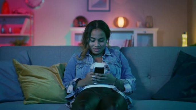美丽微笑的年轻黑人女孩坐在沙发上，在智能手机上聊天。她开朗快乐，看着电视屏幕一秒钟。舒适的房间被温暖
