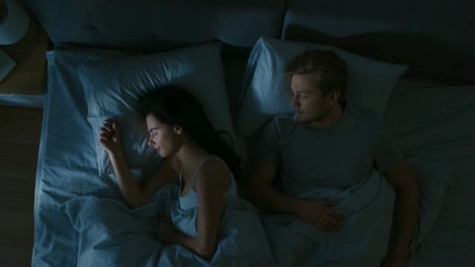 晚上的俯视图床: 迷人的年轻夫妇睡在一起，互相拥抱。蓝色夜间颜色，寒冷微弱的灯柱光线透过窗户照射。