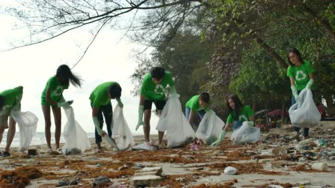 一群穿着绿色t恤的亚洲年轻人志愿者用装满垃圾的塑料袋清理海滩。安全生态概念。4k分辨率。