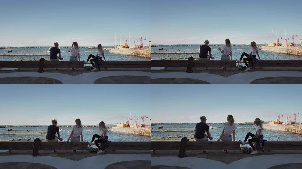 两个漂亮的女孩和英俊的男孩坐在码头的长凳上，欣赏海景港口。在后台的工业仓库和时尚的潮人区