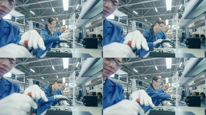 穿着蓝色工作服和防护眼镜的女电子工厂工人正在用螺丝刀组装笔记本电脑的主板。拥有多名员工的高科技工厂设
