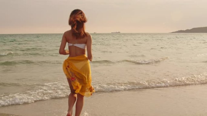 在沙滩上跑步的亚洲女人。年轻快乐的女性在比基尼放松和有趣的热带海时，日落而度假，夏季旅行的概念。慢动