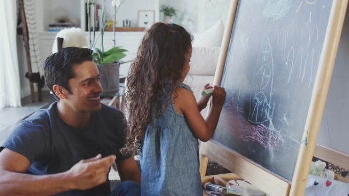 父亲坐在家里的地板上，和他的小女儿在黑板上画画，特写