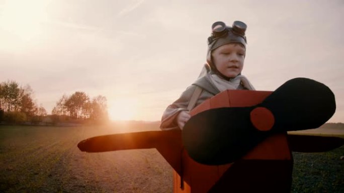 快乐的小男孩穿着有趣的纸板飞机服装沿着日落秋天的田野奔跑，玩航空飞行员慢动作。