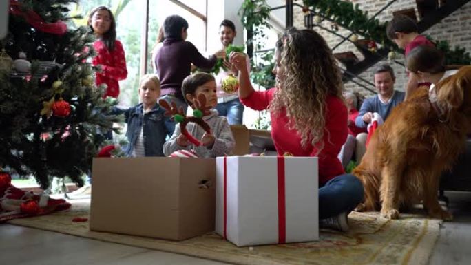 美丽的年轻母亲和儿子从盒子里拿出圣诞装饰品，而家人正在聊天，并在背景中进行有趣的装饰
