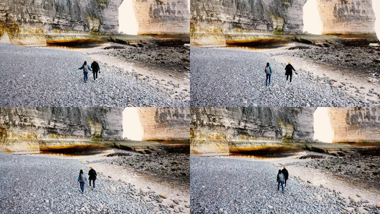 无人机摄像机跟随快乐的旅游男女沿着美丽的大圆石滩走到诺曼底雄伟的岩石。