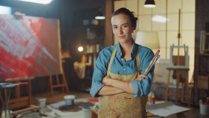 才华横溢的年轻女艺术家的肖像被油漆弄脏，穿着围裙，拿着油漆刷微笑着看着相机。地道的创意工作室，周围放
