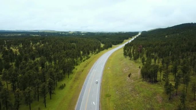 无人驾驶飞机飞越空旷的高速公路，在美丽的野生绿树森林山丘中间，白色汽车在行驶。