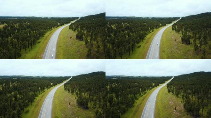 无人驾驶飞机飞越空旷的高速公路，在美丽的野生绿树森林山丘中间，白色汽车在行驶。