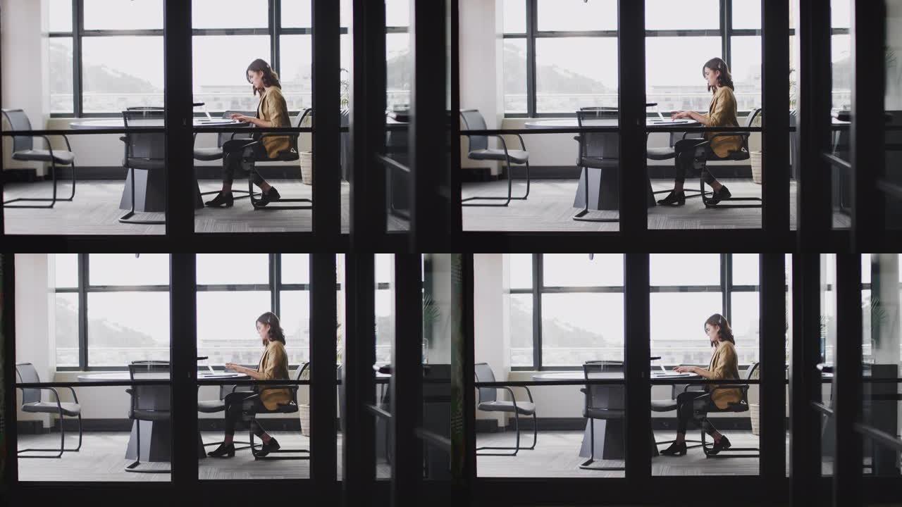 年轻的白人女商人独自在办公室工作，透过玻璃墙看到，全长，缩小