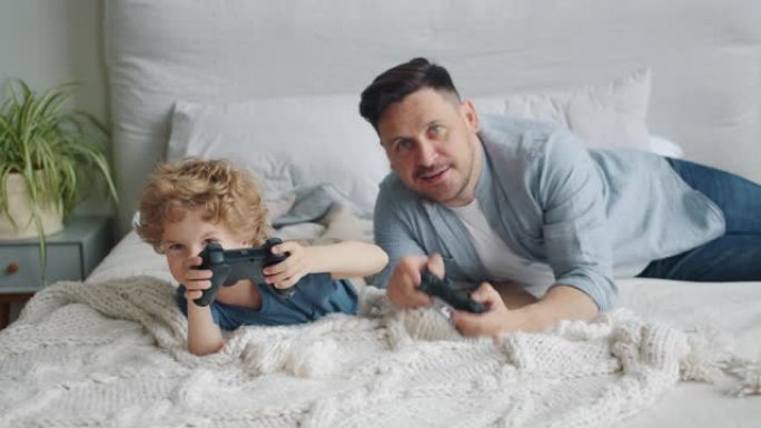 父亲和孩子在家里的床上玩电子游戏按下操纵杆上的按钮