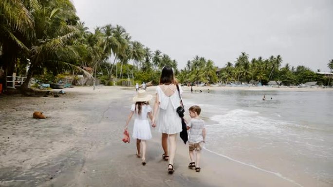 相机跟随快乐的年轻母亲和相机一起散步，两个孩子在拥挤的海洋海滩上享受假期。