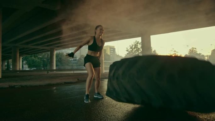 美丽自信的健身女孩穿着黑色运动上衣在街上做运动。她在城市环境中翻转一个大轮胎。