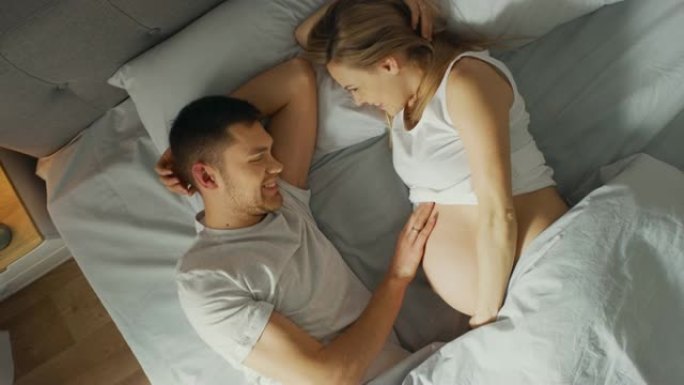 幸福的年轻夫妇在床上拥抱在一起，年轻的女人怀孕了，充满爱心的丈夫温柔地抚摸着她的肚子。顶视图摄像机。