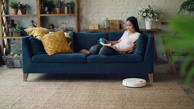 当机器人胡佛正在打扫地板做家务时，放松的亚洲女人正坐在家里的沙发上看书翻页。休闲和技术概念。