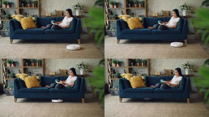 当机器人胡佛正在打扫地板做家务时，放松的亚洲女人正坐在家里的沙发上看书翻页。休闲和技术概念。