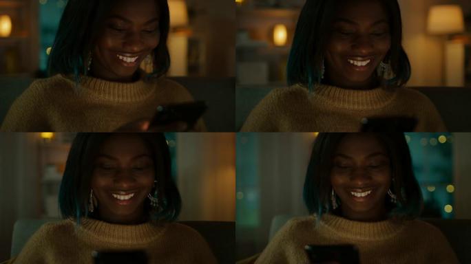 美丽的黑人女孩的肖像坐在家里的沙发上时使用智能手机，她的脸被屏幕灯照亮。在可爱的女人微笑和浏览通过互