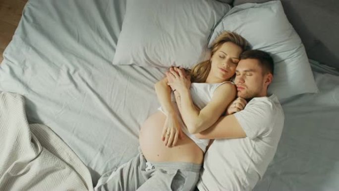 幸福的年轻夫妇一起睡在床上，可爱的年轻人在睡觉时互相抱着。美丽的女人怀孕了，有爱心的伴侣支持她。