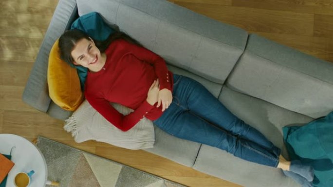 穿着红色套头衫和蓝色牛仔裤的年轻女孩躺在带枕头的沙发上。看着上面微笑。舒适的客厅，配有现代室内地毯、