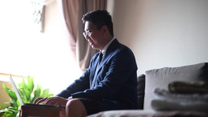 一位打着领带、穿着西装、穿着短裤的亚洲华人中年男子坐在客厅的沙发上，用笔记本电脑与他的商业伙伴和同事