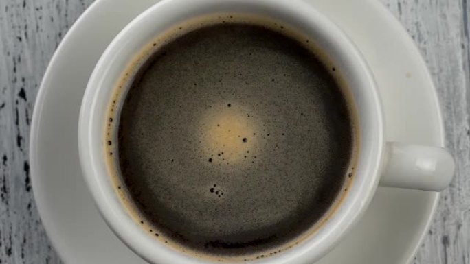咖啡泡沫在装有咖啡的白色杯子中旋转。破旧的白色木质背景。自上而下的视图。