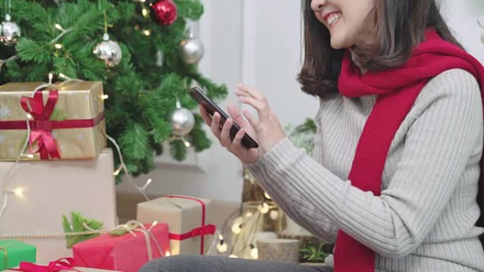 年轻迷人的亚洲女性使用智能手机与她的朋友在客厅聊天，对圣诞树背景感到满意。