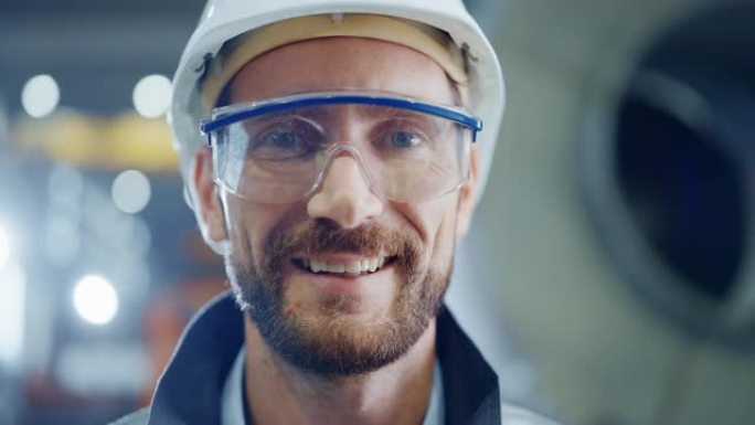 微笑的专业重工业工程师/工人穿着安全服，护目镜和安全帽的肖像。在背景中没有聚焦的大型工业工厂