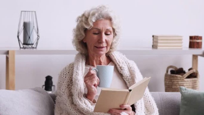 微笑的老妇人看书喝茶坐在沙发上