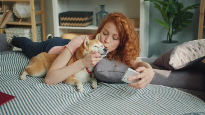 可爱的女人亲吻可爱的小狗，用智能手机在沙发上自拍