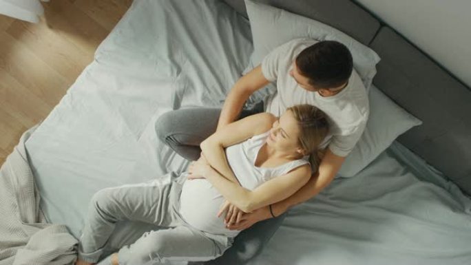 幸福的年轻夫妇在床上拥抱在一起，年轻的女人怀孕了，充满爱心的丈夫拥抱着她，温柔地爱抚着她的肚子