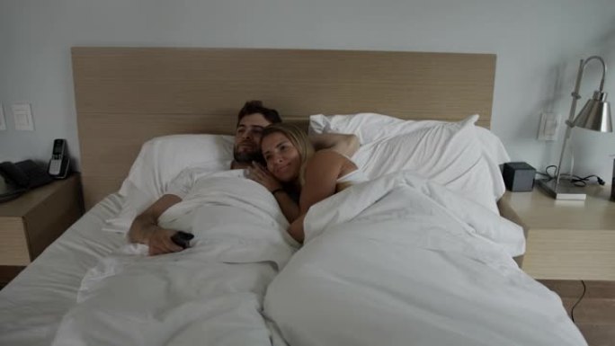 年轻夫妇躺在床上拥抱在一起看电视