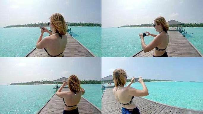 女士在马尔代夫热带蓝海上空的阳光码头上使用照相手机