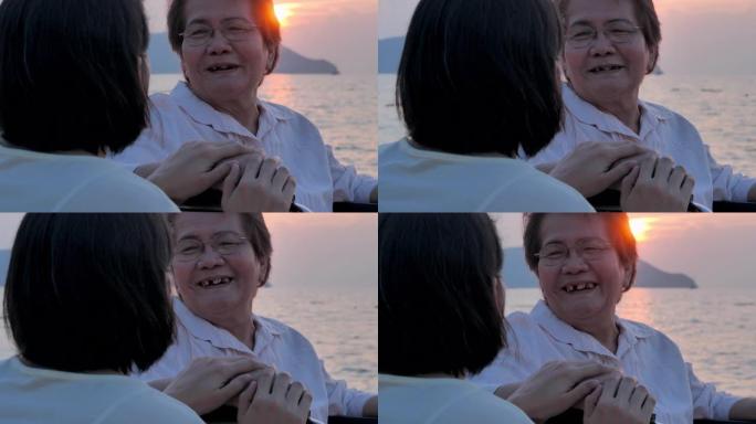 微笑的亚洲老年母亲和成年女儿在海滩上拥抱的肖像。快乐的女儿在一起度过时光时牵着照顾者。高级假期，假期