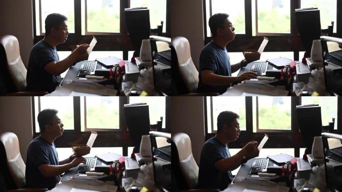一个亚洲华人中年男子正在吃午餐，在他的家庭办公室工作的时候吃他的午餐
