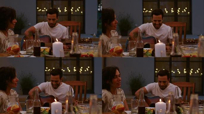 坐在餐桌上的英俊年轻人在夏季晚上的花园聚会庆典上弹吉他。朋友在浪漫的气氛中聆听动人的音乐。