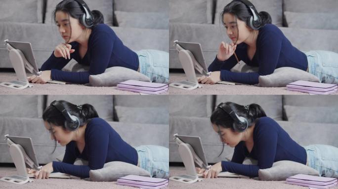 迷人的亚洲少女戴着耳机和老师在平板电脑屏幕上收听和学习在线家庭学校，并在笔记本冠状病毒或covid 