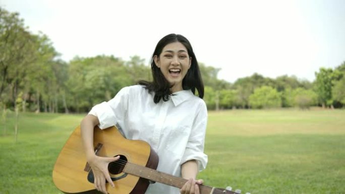 年轻迷人的亚洲女子在夏日公园弹吉他的肖像。具有幸福情感概念的女人。慢动作。