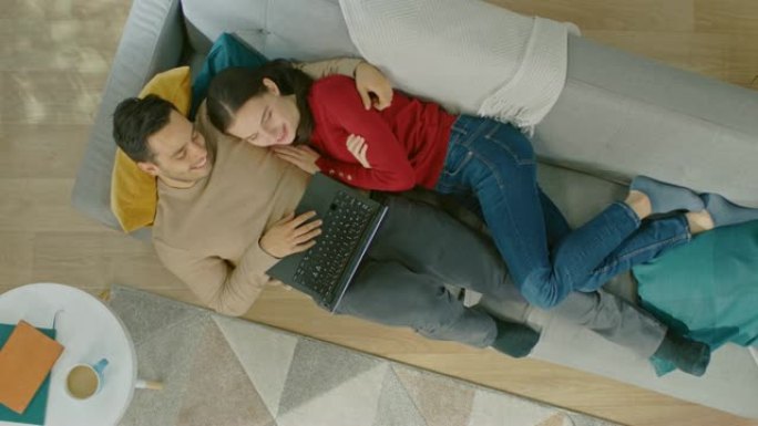 年轻美丽的夫妇躺在沙发上，用笔记本电脑。他们看着上面微笑。舒适的客厅，配有现代室内植物、桌子和木地板