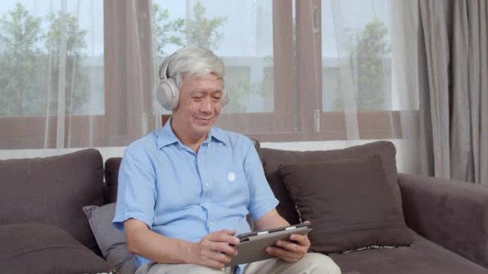 亚洲老人在家放松。亚洲老年男性快乐地戴着耳机，使用平板电脑收听播客，躺在客厅的沙发上。