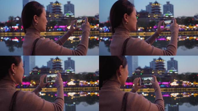 美丽的年轻亚洲女人的侧视图晚上用手机拍摄美丽的城市景色美丽的中国游客在霓虹灯下在河边的九岩桥成都欣赏