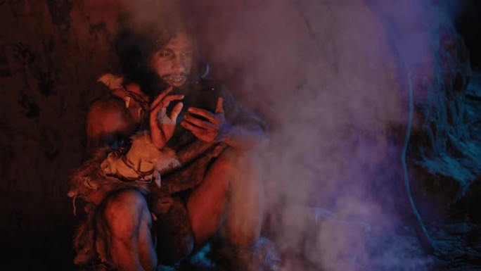 穿着动物皮的史前原始狩猎采集者部落在夜间在洞穴中使用智能手机。尼安德特人/智人男性在手机上浏览互联网