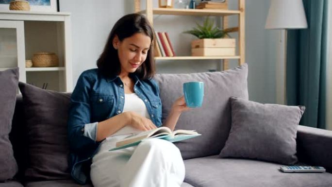 聪明的年轻女士拿着一杯茶坐在公寓的沙发上看书