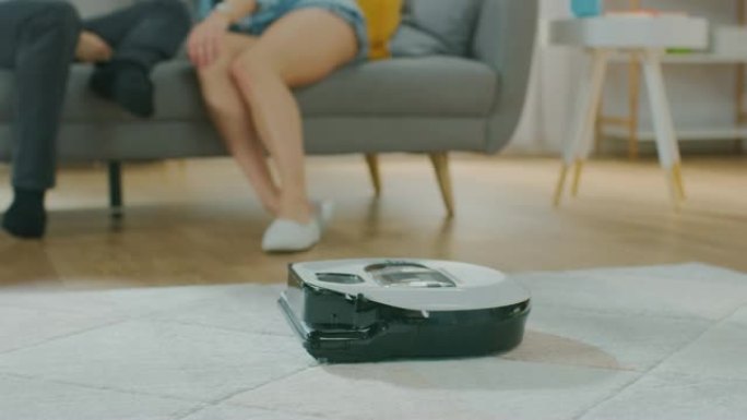智能机器人真空吸尘器从地毯上吸尘的特写镜头。美丽的夫妇坐在沙发上，在后台聊天。科技家电设备超越了它们